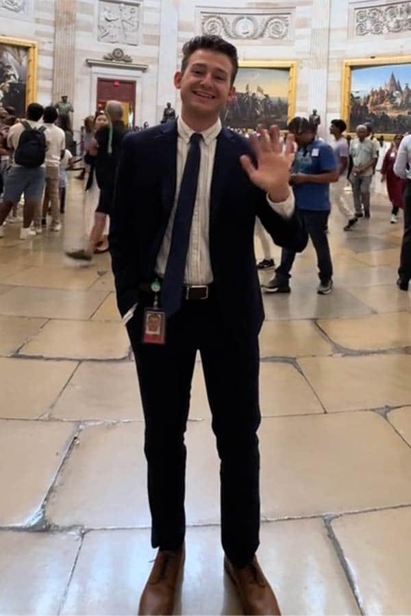 Peyton Willis, a male student wearing a suit waves from inside the U.S. Capitol. Other Capitol tourists and visitors can be seen in the background.